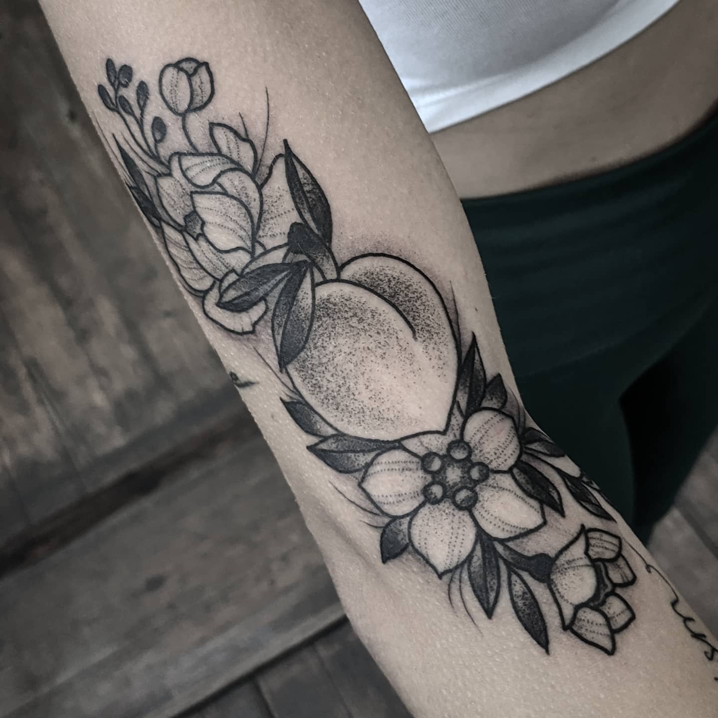 A detailed black and grey peach tattoo with florals on a forearm
